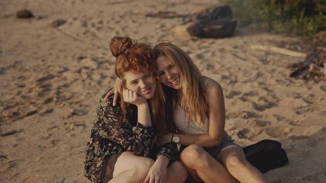 Redhead and blonde hipster girls sitting on the beach and smiling. Pretty female kissing her girlfriend during sunset. Friends hugging, concept of isolation together, vacation, friendship