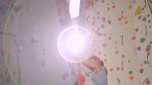 SLOW MOTION, CLOSE UP, LENS FLARE: Athletic girl reaches up to grip a crimp hold after looping her belay rope into a carabiner. Bright sun rays shine on female rock climber lead climbing indoors.