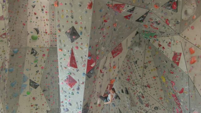 AERIAL: Athletic Caucasian woman climbing in a training center hangs on a rope after failing to grip a slippery sloper. Fit young female climber falls while trying to grip a jug hold on a red volume.