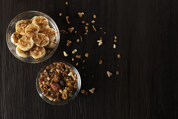 Bananas cut into slices with granola and cereal on dark gray wooden background in glass bowls on a table. Top view, copy space
