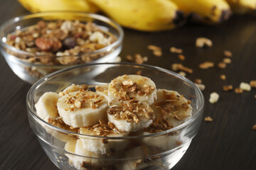 A banquet of bananas with granola and cereal on dark gray wooden background and a sliced banana in a glass bowl on a table. Selective focus