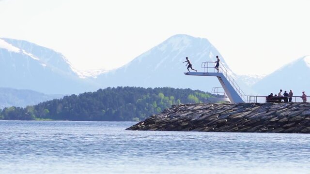 Slow motion shot of a person jumping off of a diving board into the sea, water splashing up. Beautiful scenic mountain view in the background. Molde, Norway.