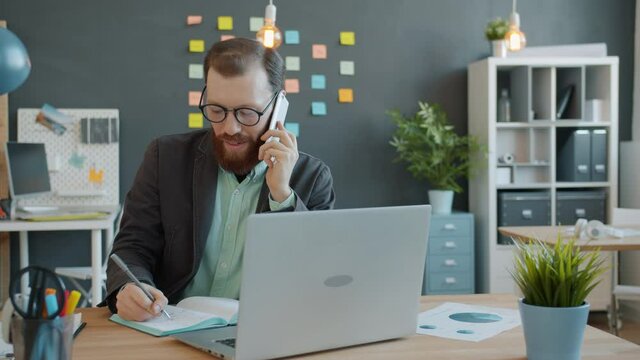 Cheerful guy is working in office making mobile phone call and using laptop taking notes in notebook. Business communication and businesspeople concept.