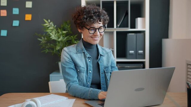 Cheerful girl busy with laptop work in office typing and looking at screen smiling enjoying indoor job in creative workplace. Technology and business concept.