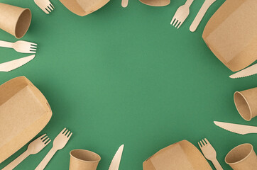 Frame of disposable paper cups and plate with wooden forks and knives on green background. Pattern...