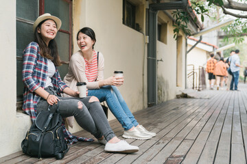 two asian chinese female friends sitting at windowsill and holding paper cups of coffee outdoor in old town. full length young girls laughing and chatting while visit little village and relax.