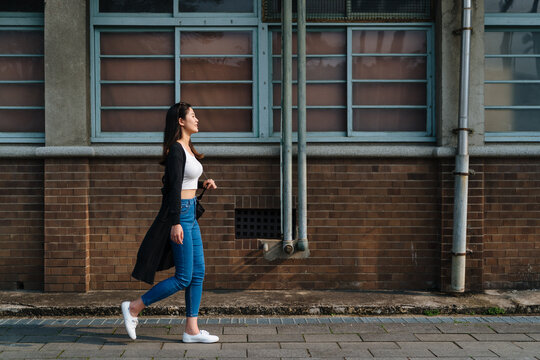 Attractive Asian Woman With Cute Smile In Stylish Crop Top In Fashion Blue Jeans Walking On Street In Old City Near Vintage Red Brick Wall. Side View Full Length Beautiful Girl Relax Enjoy Sunshine