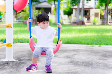 Cute baby boy wearing a white shirt is sitting on a swing on the playground. Asian children sitting alone. The background is a fresh green lawn. Child 2 years old.