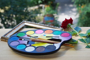 A palette with watercolor paints, brushes and a red rose on a windowsill.