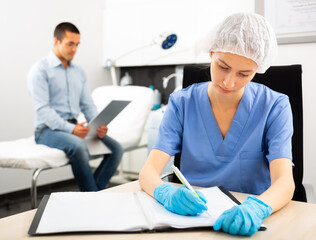 Obraz premium Portrait of young woman beautician sitting at desk writing notes while man patient checking result of procedure looking in mirror