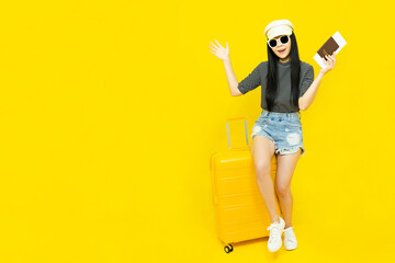 Young Asian female tourist in casual clothes and sunglasses Carrying a passport to go to travel sitting on a luggage isolated on yellow background copy space,Vacation,holidays and travel concept