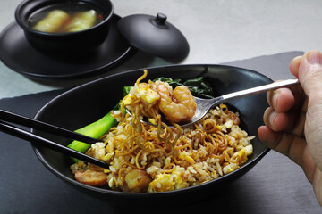 fried rice and instant noodles with shrimps and cantonese kale in the black bowl served on the black plate together with vegetable soup on the grey table in the restaurant