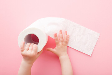 Baby girl hands holding soft, white toilet paper roll on light pink table background. Pastel color....