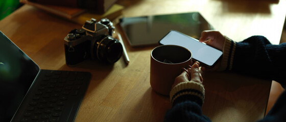 Hand using mock up smartphone and holding mug on rustic worktable with camera and  tablet