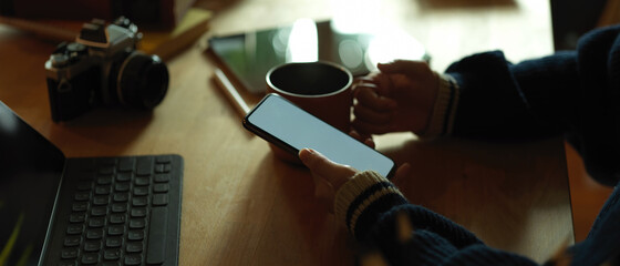 Hand using mock up smartphone and holding mug on rustic worktable with camera and  tablet
