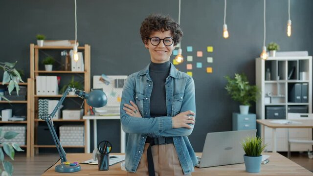Portrait of attractive girl with dark curly hair standing in office smiling looking at camera wearing glasses and trendy clothing. Youth and workplace concept.