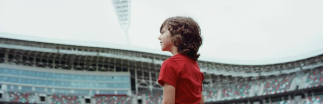 Cute little kid boy soccer player standing on an empty stadium, dreaming of becoming professional player, soccer star. RED cinema camera RAW graded footage