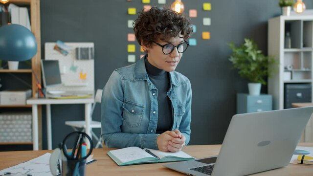 Businesswoman is alking online making video call with laptop from creative office speaking and gesturing sitting at desk alone. Youth and communication concept.