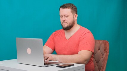 Young happy businessman in a pink t-shirt works on a laptop on a green screen. Portrait of a male talking man looking into his laptop. Man working at his desk in the office.