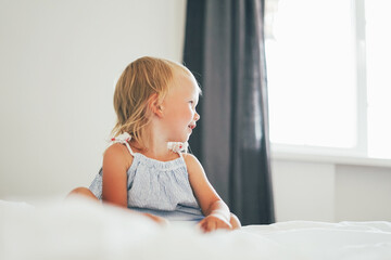 Fototapeta premium Cute blonde baby girl on bed with white bed linen at the bright interior