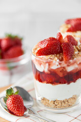 Homemade layered dessert with fresh strawberries, cream cheese or yogurt, granola and strawberry jam in glasses on white wood background. Healthy organic breakfast or snack concept. Selective focus.