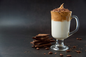 Glass of trendy fluffy creamy dalgona or whipped instant coffee with milk, coffee beans and chocolate on the dark background. New popular food and drink trend concept. Copy space.