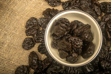 Dried prunes in a metal bowl and dried prunes scattered on a background of coarse-textured burlap. Close up.