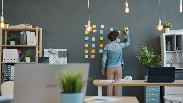 Back view of female office worker writing on colorful sticky notes planning business project focused on creative activity. Ideas, millennials and entrepreneurship concept.