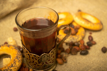 tea, driedA faceted glass of tea in a vintage Cup holder, dried rosehip fruit and small bagels on a background of homespun fabric with a rough texture. Close up. rosehip fruit