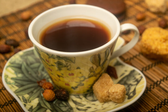 A Cup of tea, dried rosehip fruit, pieces of brown cane sugar, and cookies on a reed Mat. Close up.
