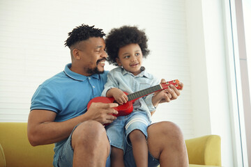 An African American father and little son singing and playing ukulele together. Dad teaching his boy play a small red ukulele on sofa at home