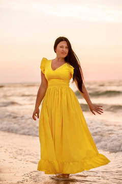 A girl in a long dress with long black hair walks at dawn along a deserted beach along the line of the surf. 