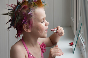 Obraz premium A little girl is getting ready for a party. A child paints her lips in front of a mirror. The girl is dressed in an evening pink dress.
