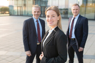 Positive successful business woman wearing office suit, standing outdoors and looking at camera. Male business colleagues standing behind. Business team concept