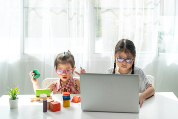 Asian girl using desktop computer for online study homeschooling during her younger sister play wooden block toy at her home. coronavirus, homeschooling or education technology concept