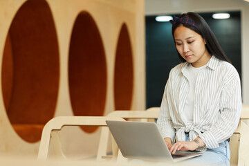 Focused Asian student girl in checkered headscarf sitting in modern lobby and writing coursework on...