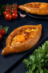 Adjarian Khachapuri – Georgian cheese bread with tomatoes, parsley and garlic on black board. Baked open pie with cheese and egg yolk filling.