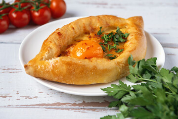 White plate with Adjarian Khachapuri – Georgian cheese bread decorated with tomatoes and parsley on white Provence style background. Baked open pie with cheese and yolk filling.