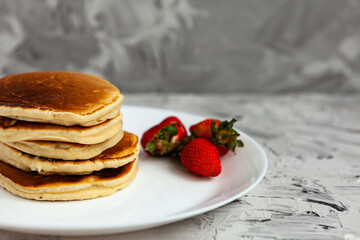 A stack of pancakes with a pink tea mug and fresh strawberries on a white plate on a light background. Horizontal orientation. Homemade american pancakes. Copy space.