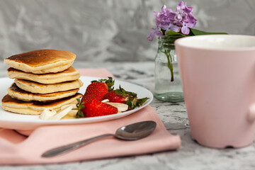 A stack of pancakes with a pink tea mug and  fresh strawberries on a white plate on a light background. A small bouquet of lilacs in a glass vase stands next to the Breakfast.
