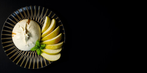 Traditional Italian, fresh, soft white cheese burrata ball of mozzarella and cream on a round dark plate, with slices of pear on a dark background. Copy space.
