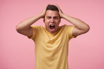 Fototapeta na wymiar Stressed young brown-eyed short haired male clutching his head with raised hands and shouting angrily with wide mouth opened, standing over pink background