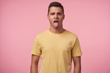 Studio shot of young pretty brown haired male sticking out his tongue while looking at camera, keeping hands along body while posing over pink background