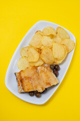 fried cod fish with olives and potato chips on white dish on yellow background