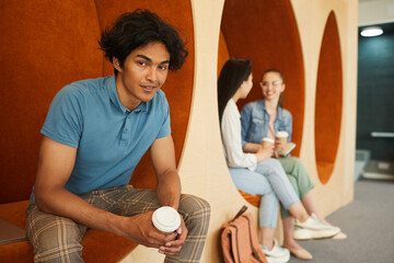Fototapeta premium Portrait of smiling handsome dark-skinned boy with disheveled hair sitting in campus lobby and drinking coffee