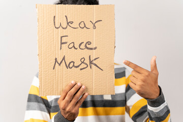 Young man holding a cardboard sign by covering his face, saying wear face mask meassage on isolated background to protect from coronavirus or covid-19.