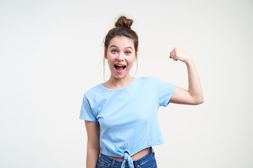 Excited young attractive dark haired woman with bun hairstyle keeping her hand raised while demonstrating her strong biceps, isolated over white background