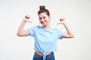 Fototapeta na wymiar Pleased young lovely brunette female dressed in casual clothes smiling positively while thumbing on herself with raised hands, isolated over white background