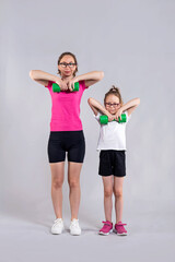 Stylish mom and daughter stand on a gray background in sportswear and with dumbbells.