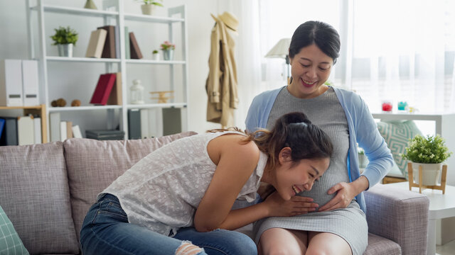 curious asian japanese woman listening to beautiful pregnant friend tummy and smiling. young lady putting ear on abdomen and careful hearing unborn baby inside sister belly on sofa. motherhood expect